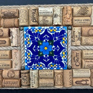 May include: A square cork board with a blue and white tile in the center. The cork board is decorated with wine corks, some with labels, and rope.