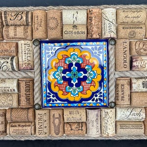 May include: A square cork board with a colorful tile in the center. The tile has a blue, yellow, red, and green floral design. The cork board is surrounded by a rope border and is filled with wine corks. Some of the corks have text on them, including "Clos du Bois", "Folie a Deive", "Woodbridge", "L'Angolo", "Beaune France", "Kendall Jackson", and "Santa Margherita".