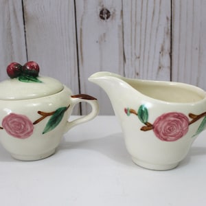 Puede incluir: Un juego vintage de azucarero y jarra de crema de color crema con un diseño floral. El juego presenta rosas rosas, hojas verdes y ramas marrones. El azucarero tiene una tapa con dos bayas rojas en la parte superior.