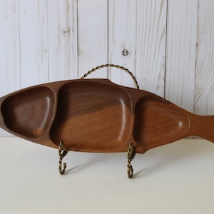 Puede incluir: Una bandeja de servir de madera con forma de pez con tres compartimentos separados. La bandeja tiene un asa y un soporte de metal dorado.