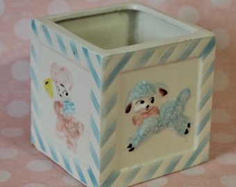 May include: A white ceramic planter with blue and white stripes. The planter features a pink lamb with a yellow bell and a blue lamb with a pink bow.