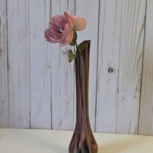 May include: A single pink flower in a tall, clear glass vase with swirling purple and green stripes. The vase has a bulbous base.