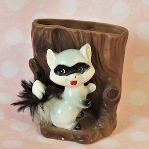 May include: A ceramic raccoon figurine with a fluffy tail, peeking out from a brown tree trunk planter. The raccoon has black eyes and a red mouth.