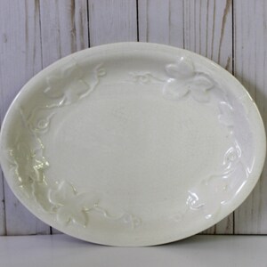 May include: Off-white oval serving platter with embossed leaf and vine design. The ceramic dish has a slightly raised rim and a smooth, glossy surface. The decorative elements are arranged around the edge of the platter.
