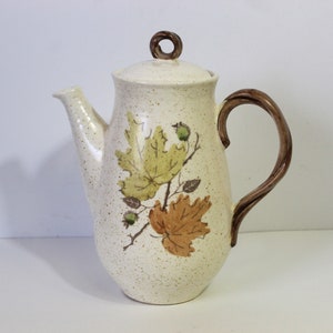 May include: A white ceramic coffee pot with a brown handle and lid. The pot has a speckled glaze and a design of brown and green leaves.