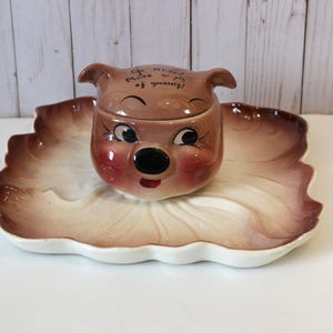 May include: A vintage ceramic pig-shaped dish set. The lid of the dish features a pig face with rosy cheeks and the text "Go ahead, make a pig of yourself!". The dish is set on a leaf-shaped platter with a white and brown color scheme.