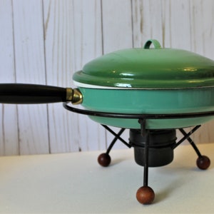 May include: A vintage green enamelware pot with a black handle and lid, sitting on a black metal stand with wooden legs.