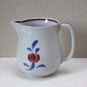 May include: A white ceramic pitcher with a red rim and handle. The pitcher is decorated with a red and blue floral design. The pitcher is likely used for serving beverages.