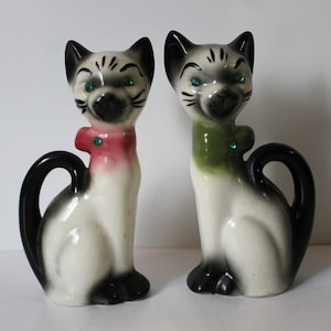 Könnte beinhalten: Zwei Keramik-Siamesen-Katzenfiguren mit schwarzen und weißen Markierungen. Die Katzen tragen grüne und rosa Halsbänder mit Juwelen-Akzenten.