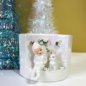 May include: A white ceramic planter with an iridescent finish, featuring a seated figure and a rabbit. The planter is decorated with small flowers. Two tinsel Christmas trees are in the background.