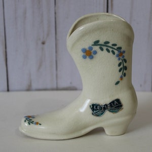 Könnte beinhalten: Eine weiße Keramikvase in Form eines Stiefels mit blauen und grünen Blumendesigns und einem Etikett mit der Aufschrift "Letizia" auf einem grün-weißen Band.