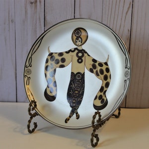 May include: A white ceramic plate with a gold rim features a black and gold illustration of a woman in a stylized Art Deco design. The plate is displayed on a gold metal stand.