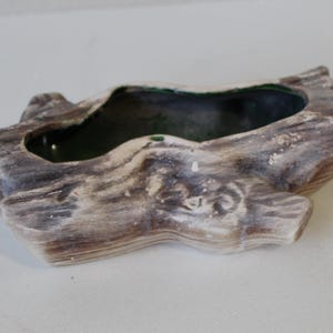 May include: A ceramic planter shaped like a weathered log. The planter has a dark interior and is painted in shades of brown, gray, and white to mimic the appearance of wood. The planter is designed to hold small plants or succulents.