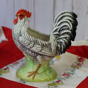 Peut inclure: Un pot de fleurs en céramique en forme de coq blanc et noir avec une crête et des barbillons rouges. Le coq est debout sur une base verte avec une patte brune.