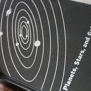 May include: A vintage book titled "Planets, Stars, and Galaxies" with a black cover. The cover features a white illustration of a solar system. The book is held by a hand.
