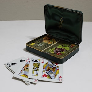 May include: A dark green rectangular storage case with a hinged lid, containing two decks of playing cards with fruit illustrations on the backs. Four playing cards are fanned out in front, including the four of clubs, king of diamonds, king of spades, and queen of hearts.