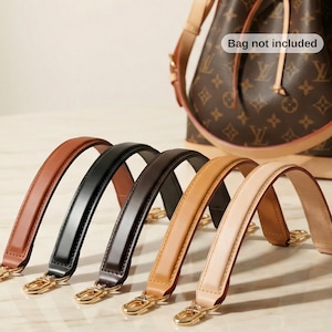 Può includere: Cinque manici di ricambio in pelle per borse in vari colori: marrone, nero, marrone scuro, beige e beige chiaro. Ogni manico ha finiture color oro. Sullo sfondo, una borsa marrone con chiusura a coulisse. Il testo "Bag not included" è visibile.