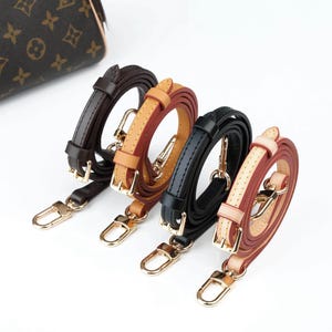 May include: Four leather purse straps in brown, tan, black, and a combination of tan and burgundy. Each strap has gold-colored metal clasps and hardware. The straps are arranged in a row against a white background.
