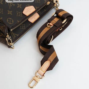 Puede incluir: Correa de bolso a rayas marrones y beige con herrajes dorados. La correa está sujeta a un bolso marrón con monograma. La imagen incluye el texto "Bag not included".