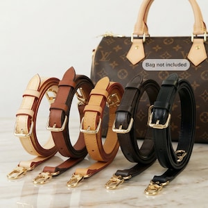 Può includere: Una collezione di tracolle per borse in pelle in vari colori: crema, marrone, beige, marrone scuro e nero. Ogni cinturino presenta elementi metallici color oro, tra cui fibbie e chiusure. Sullo sfondo, una borsa marrone fantasia, con la scritta "Bag not included".