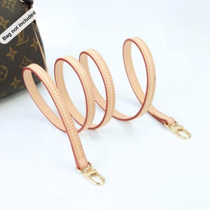May include: A light tan leather purse strap with red edging and gold-colored clasps. The strap is shown in a looping pattern against a white background. The bag is not included.