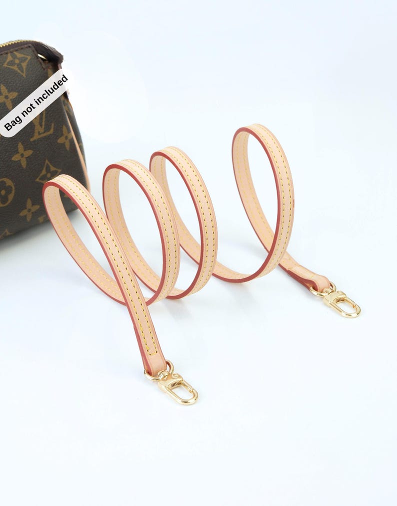 May include: A light tan leather purse strap with red edging and gold-colored clasps. The strap is shown in a looping pattern against a white background. The bag is not included.