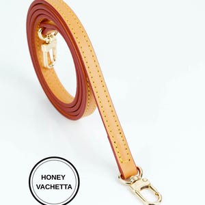 May include: A honey vachetta leather strap with gold-tone hardware. The strap is a light tan color with a reddish-brown interior and yellow stitching. The strap is coiled and has a gold-tone clasp. The text "HONEY VACHETTA" is in a black circle.