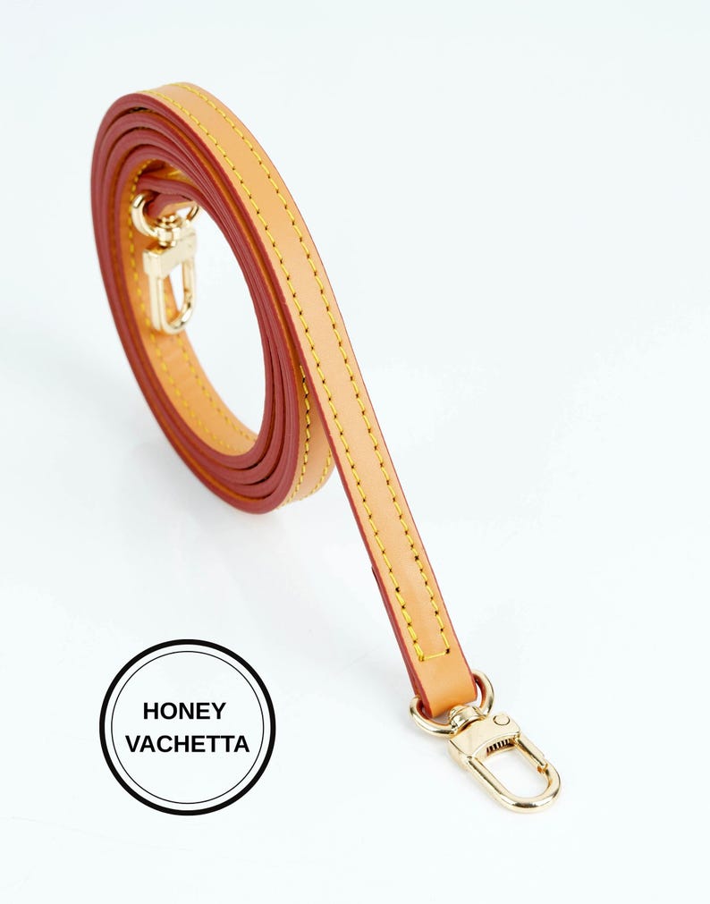 May include: A honey vachetta leather strap with gold-tone hardware. The strap is a light tan color with a reddish-brown interior and yellow stitching. The strap is coiled and has a gold-tone clasp. The text "HONEY VACHETTA" is in a black circle.