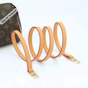 May include: A light tan leather purse strap with gold-colored clasps. The strap is coiled and has a red interior with yellow stitching. A brown purse with a monogram pattern is visible in the background.
