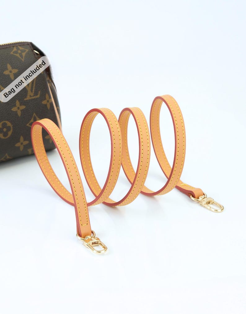 May include: A light tan leather purse strap with gold-colored clasps. The strap is coiled and has a red interior with yellow stitching. A brown purse with a monogram pattern is visible in the background.