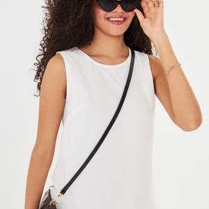 May include: A woman wearing black cat-eye sunglasses and a white sleeveless dress. A black strap crosses the body, holding a brown purse with a logo print. A gold bracelet and ring are also visible.