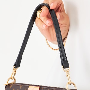 May include: Brown and tan handbag with a black strap and gold-colored hardware. The bag features a repeating pattern of the brand's logo. The strap is held by a hand. The text "Bag not included" is visible.