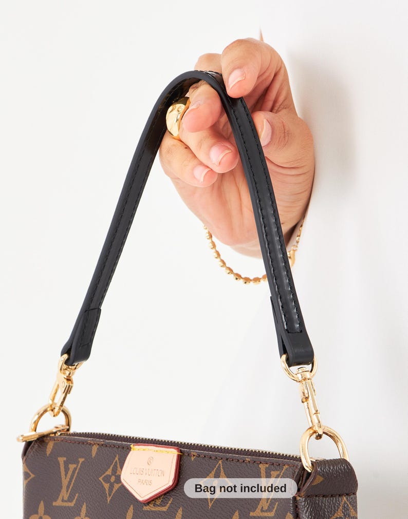 May include: Brown and tan handbag with a black strap and gold-colored hardware. The bag features a repeating pattern of the brand's logo. The strap is held by a hand. The text "Bag not included" is visible.
