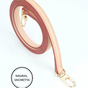 May include: A rolled, light pink and beige leather bag strap with gold-colored metal clasps. The strap is stitched with yellow thread and features the text "NAURAL VACHETTA" in a black circle.