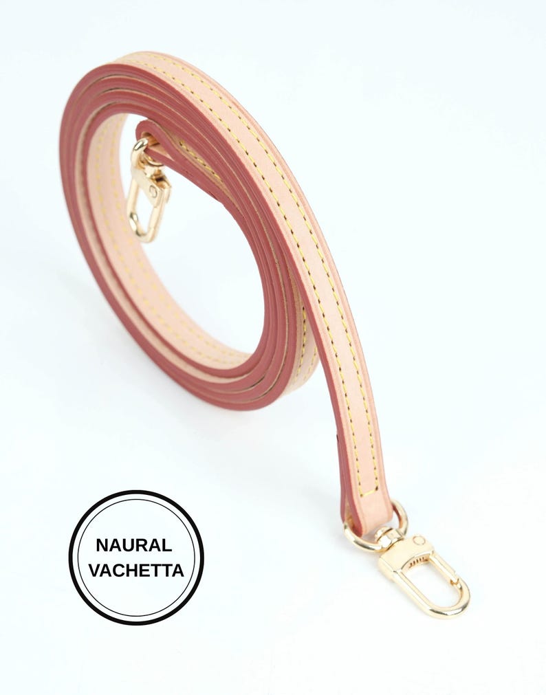May include: A rolled, light pink and beige leather bag strap with gold-colored metal clasps. The strap is stitched with yellow thread and features the text "NAURAL VACHETTA" in a black circle.