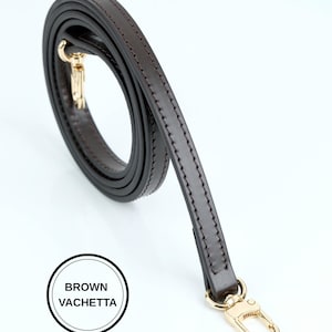 May include: A brown leather bag strap with gold-colored hardware. The strap is a dark brown color and appears to be made of high-quality leather. The text "BROWN VACHETTA" is in a white circle.