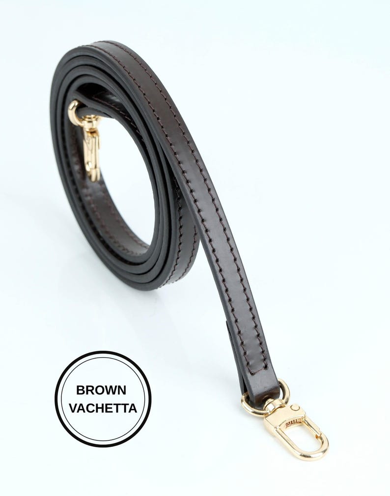 May include: A brown leather bag strap with gold-colored hardware. The strap is a dark brown color and appears to be made of high-quality leather. The text "BROWN VACHETTA" is in a white circle.