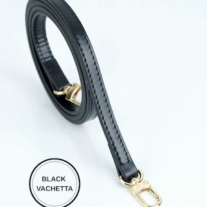 May include: A black leather strap with gold-colored hardware. The strap is made of black vachetta leather and has a stitched design along the edges. The strap is curled and has a gold-colored clasp.