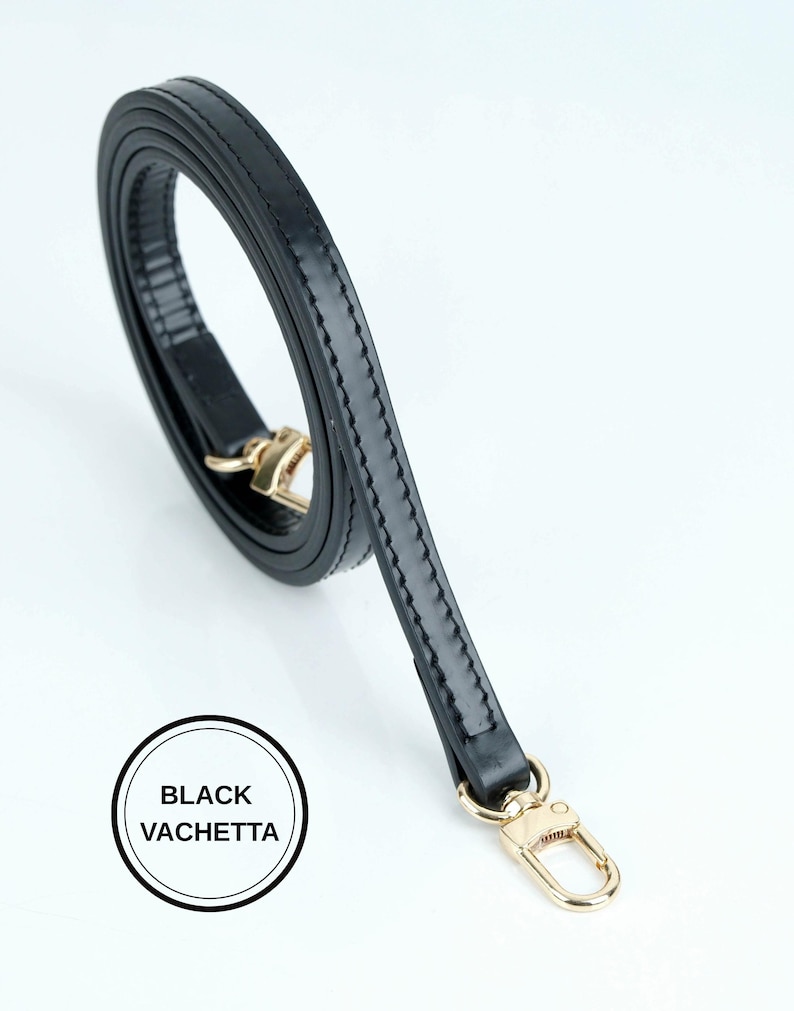 May include: A black leather strap with gold-colored hardware. The strap is made of black vachetta leather and has a stitched design along the edges. The strap is curled and has a gold-colored clasp.