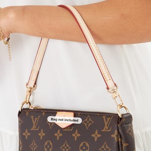 May include: Brown monogrammed handbag with a cream-colored strap and gold-tone hardware. The bag has a zipper closure and a small tag that reads "Bag not included". The strap is attached with gold clasps.