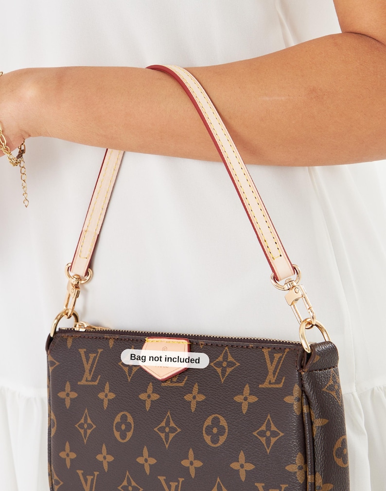 May include: Brown monogrammed handbag with a cream-colored strap and gold-tone hardware. The bag has a zipper closure and a small tag that reads "Bag not included". The strap is attached with gold clasps.