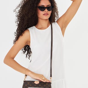 May include: A woman wearing black cat-eye sunglasses and a white dress. A brown crossbody bag with a monogram pattern is featured, with a label that reads "Bag not included". The bag has gold-colored hardware.
