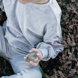 Könnte beinhalten: Eine Person trägt einen hellgrauen Cordpullover und eine passende Hose und sitzt in einem Feld mit lila Heidekraut. Sie hält einen grauen Stein in der Hand.