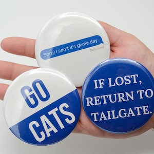 May include: Three round blue and white buttons with text. The first button says "GO CATS" in white letters on a blue background. The second button says "Sorry I can't it's game day" in a blue text bubble on a white background. The third button says "IF LOST, RETURN TO TAILGATE." in white letters on a blue background.