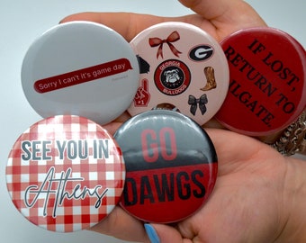 May include: Five round buttons with different designs. One button is white with red text that says "Sorry I can't it's game day". Another button is pink with a red bulldog, a red bow, a red football, a red number one, a red and white finger pointing up, a red and white bow, a red cowboy boot, and a red and white Georgia G. Another button is red with white text that says "IF LOST, RETURN TO WILGATE.". Another button is red and white checkered with black text that says "SEE YOU IN Athens". The last button is black and red with white text that says "GO DAWGS".