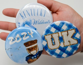 May include: Three round blue and white buttons with different designs. The first button has a white background with the text "Kentucky Wildcats" in black. The second button has a blue background with a white and blue checkered pattern and the text "UK" in white with gold glitter. The third button has a blue background with the text "UKY'S VERSION 2021" in white and a cartoon image of a wildcat.