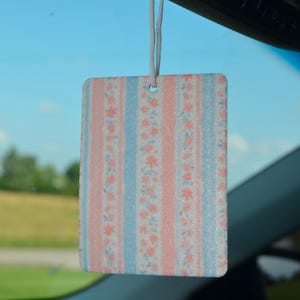 May include: Rectangular car air freshener with a floral pattern in pink and blue stripes. The air freshener is hanging from a string, visible against a blue sky background. The design features small pink flowers.