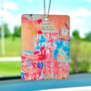 May include: A pink and blue car air freshener with the text "Jesus Loves You" in white letters. The background is a collage of colorful hearts, flowers, and other abstract designs.