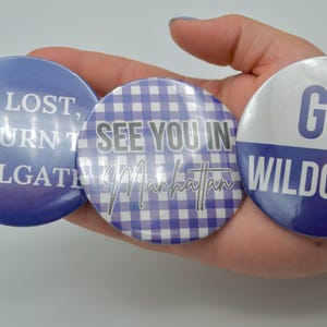 Puede incluir: Tres botones redondos azules y blancos con diferentes textos. El primer botón dice "Si se pierde, devuelva al tailgate". El segundo botón tiene un patrón de cuadros vichy y dice "Nos vemos en Manhattan". El tercer botón dice "Go Wildcats".