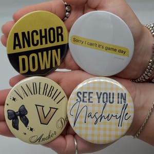 May include: Four round buttons with text and designs. One button says "ANCHOR DOWN" in black and gold. Another says "Sorry I can't it's game day". The third has a bow and the word "VANDERBILT". The last says "SEE YOU IN Nashville" on a yellow gingham background.